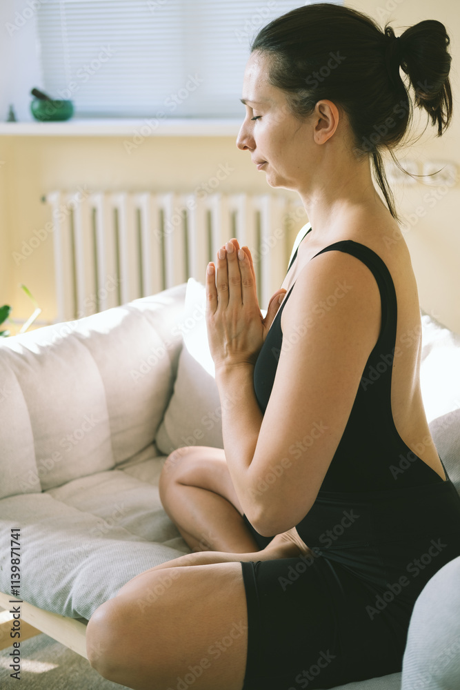 Young attractive woman practicing yoga, sitting in Padmasana on couch, exercising, Lotus pose, namaste, working out, wearing sportswear. Young attractive woman practicing yoga, sitting in Padmasana on couch, exercising, Lotus pose, namaste, working out, wearing sportswear.