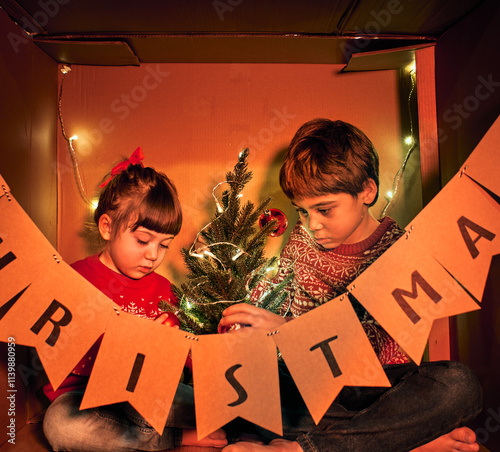 Two children in festive sweaters decorate a small Christmas tree inside a lit cardboard box. A banner spelling CHRISTMAS hangs on the box. The warm lights and decorations create a cozy holiday scene