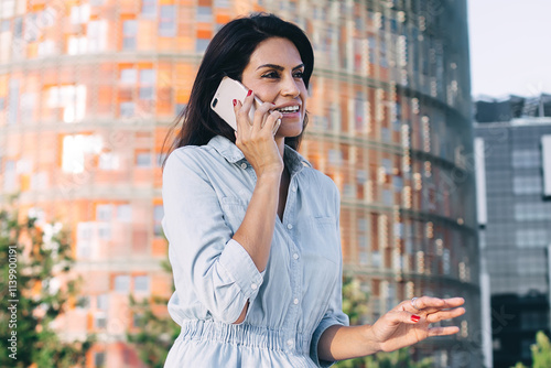 Smiling caucasian female traveler enjoying connection in roaming sitting with mobile phone on urban setting background, positive hipster girl having smartphone conversation on sunny day leisure #1139900191