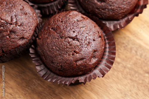 Chocolate muffins with rich texture and glossy tops. Placed in brown paper liners on a wooden surface.  photo