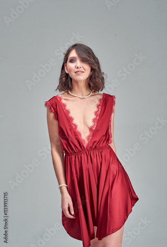 Woman in red dress poses for photo and studio. She is wearing bra, her arms are crossed. Dress is red, has flowing, elegant design.