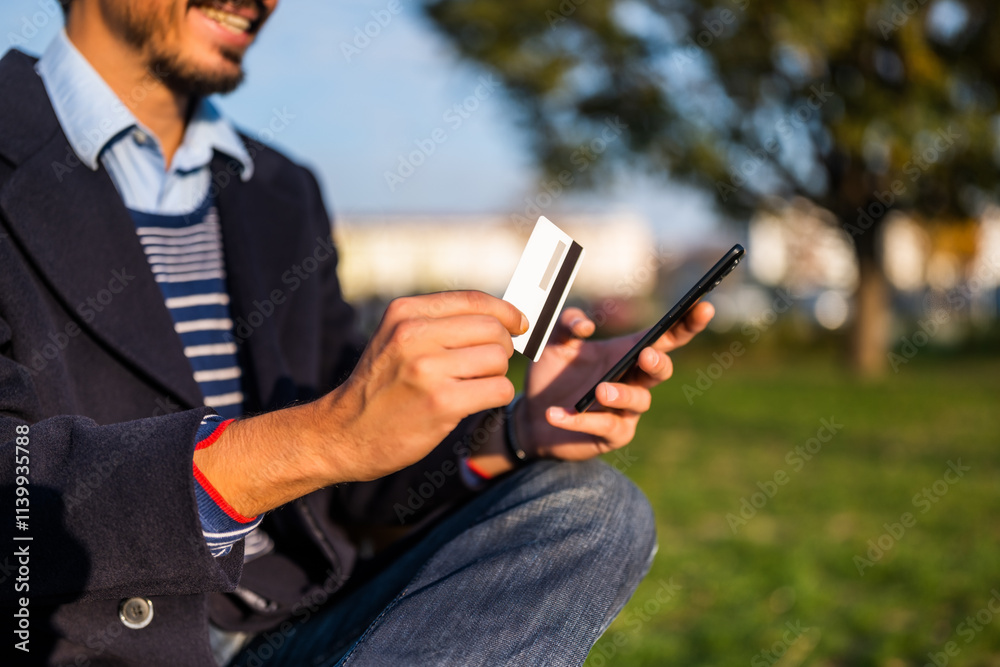 Close up photo of businessman using mobile phone and credit card while sitting on the bench in the park. Focus on credit card and phone. Close up photo of businessman using mobile phone and credit card while sitting on the bench in the park. Focus on credit card and phone.