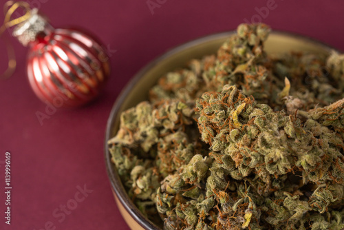 on a dark red background in a bowl some dry buds of medical marijuana, next to a Christmas ball
