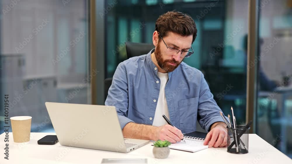 Focused businessman in glasses writing in notebook. Professional male working at modern office exuding professionalism and determination. Businessman wearing blue shirt and feeling concentrated. Focused businessman in glasses writing in notebook. Professional male working at modern office exuding professionalism and determination. Businessman wearing blue shirt and feeling concentrated.