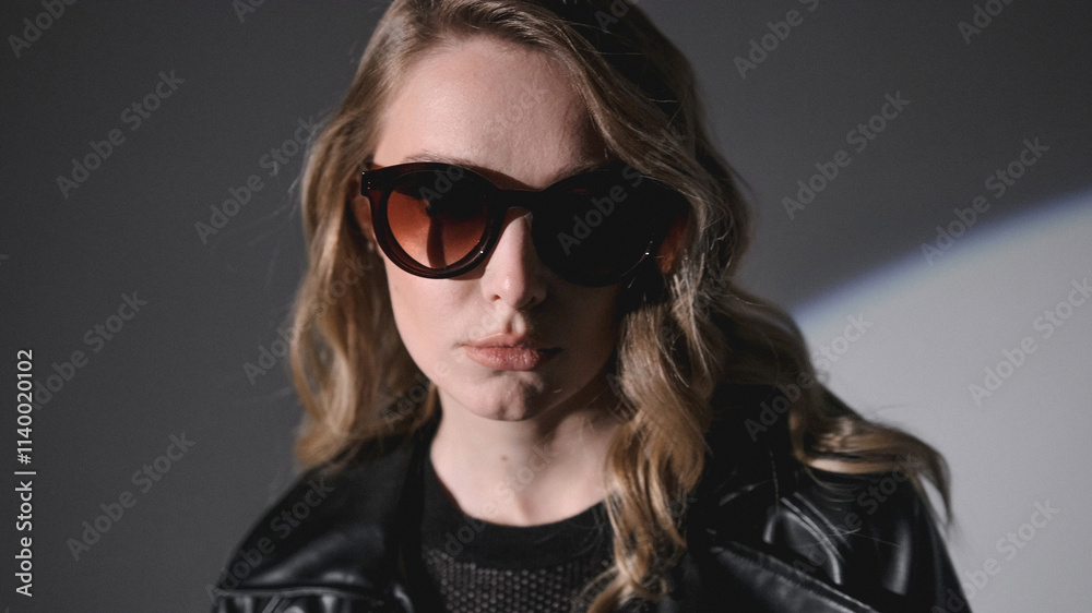 A close-up portrait of a woman with blonde wavy hair, wearing sunglasses and a black leather jacket. The lighting is dramatic, with a spotlight effect. A close-up portrait of a woman with blonde wavy hair, wearing sunglasses and a black leather jacket. The lighting is dramatic, with a spotlight effect.