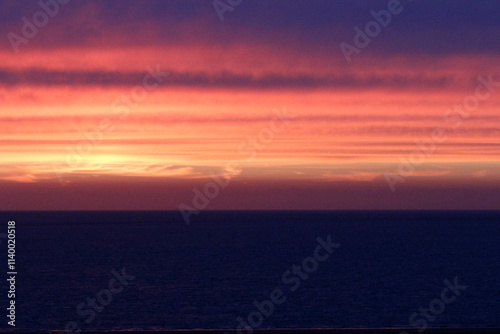 A vibrant sunset over a dark ocean.  Warm colors transition to cool as day fades.  Calm seas reflect the fiery sky.