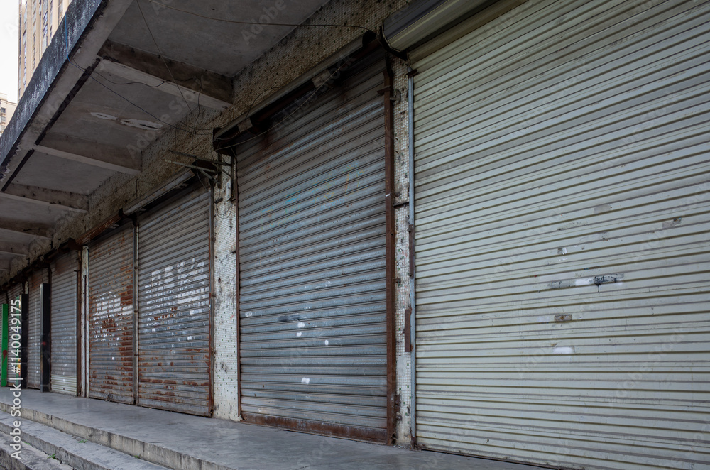 A row of closed and shuttered storefronts on a city street. The metal security gates, with signs of age and neglect, are pulled down. A sense of economic decline and a rundown commercial area in China A row of closed and shuttered storefronts on a city street. The metal security gates, with signs of age and neglect, are pulled down. A sense of economic decline and a rundown commercial area in China