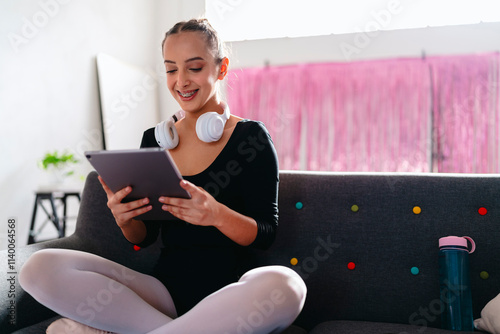 A young ballerina sits cross-legged on a gray couch, wearing white headphones and using a tablet, with a pink curtain and vibrant modern decor visible in the background. photo