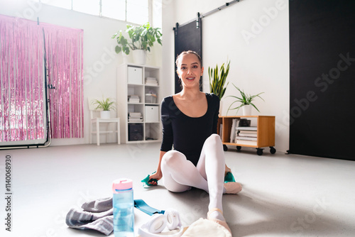 A ballerina in a black leotard and white tights practices a standing pose in a bright studio with pink shimmering curtains, a clothing rack, and neatly arranged studio decor. photo