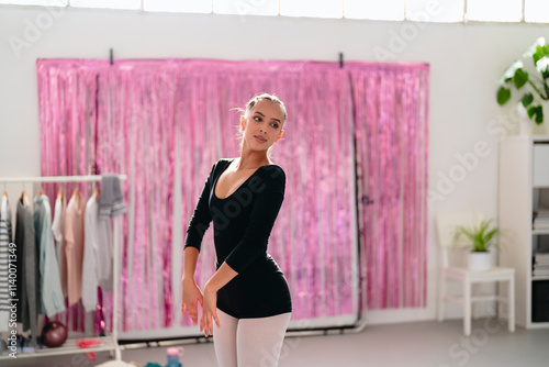 A ballerina in black leotard and white tights performs a graceful dance pose with arms extended near modern black panels, in a well-lit minimalist studio with wooden shelves and potted plants. photo