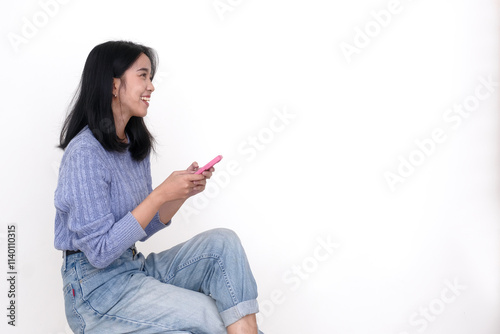 Side view: A young woman sitting on a wooden bench while holding a mobile, looking forward with a smiling, happy expression photo