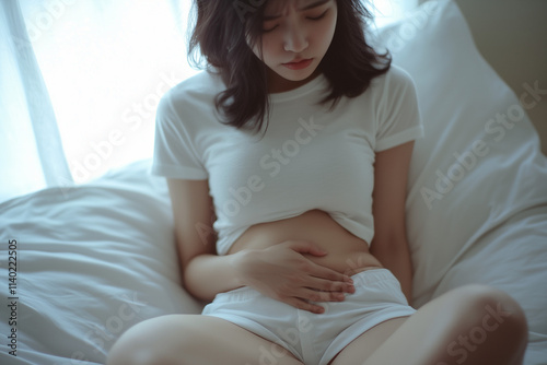 asian woman clutching belly in pain from period cramps,  endometriosis, or pregnancy sitting on bed wearing white