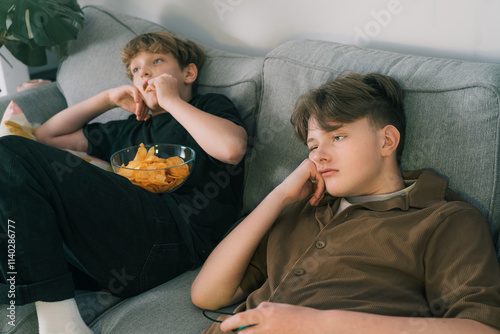 Foto Two tired teenager brothers lying on sofa looking bored, watching tv, eating snacks at home