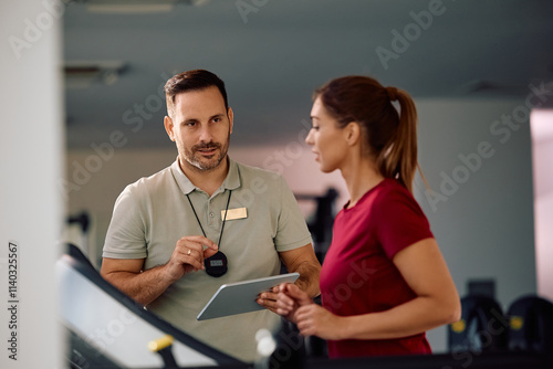 Personal trainer giving instructions to athletic woman who is running on treadmill in gym.