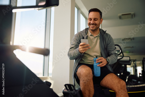 Happy athlete using mobile phone during water break in gym. photo