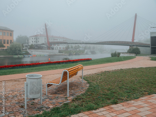 Sitting area by a river with view on city buildings and modern pedestrian bridge in Jelgava, Latvia, Town is in fog in the background. Calm surreal mood. No people. Park design. photo