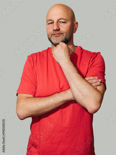 A man in a red shirt is standing with his arms crossed. He has a serious expression on his face, light color background. Male in his 40s with grey beard and bold head, slim body type. photo