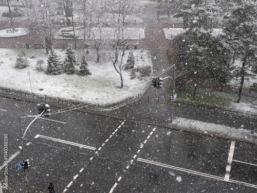 Obraz na płótnie View from the window over heavy fluffy snow falling over the pedestrian crossing with red traffic light and urban park with a lonely person walking. Snowy winter day. w salonie