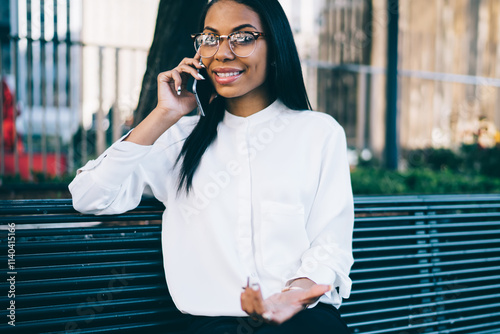 Portrait of successful african american office worker communicating with friends on telehone device resting on bench in urban setting.Positive dark skinned young woman calling on modern cellular photo