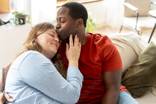 Relaxed middle aged woman hugging African American boyfriend while sitting on couch at home