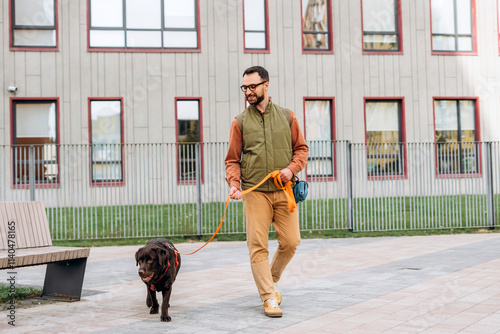 Happy Latin bearded man wearing casual clothes leading black dog, labrador on leash on the street photo