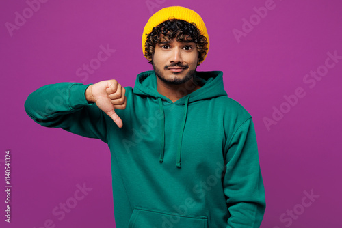 Young sad disappointed dissatisfied Indian man he wear green hoody yellow hat casual clothes showing thumb down dislike gesture isolated on plain purple background studio portrait. Lifestyle concept. photo