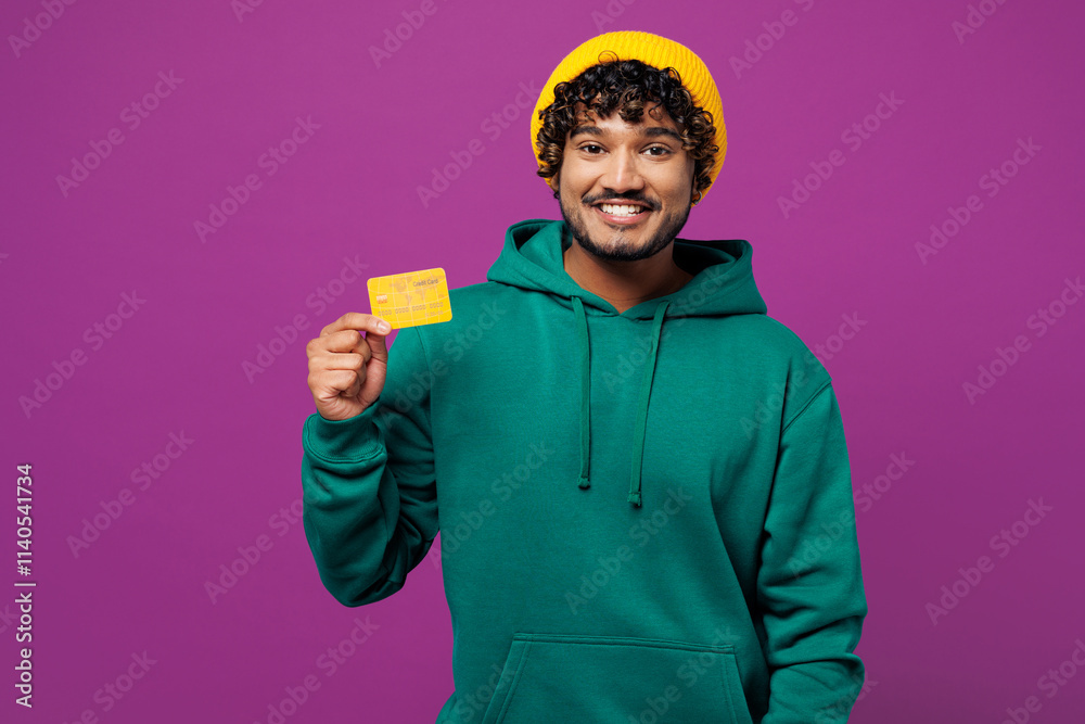 Young happy smiling cheerful Indian man he wear green hoody yellow hat casual clothes hold in hand mock up of credit bank card isolated on plain purple background studio portrait. Lifestyle concept. Young happy smiling cheerful Indian man he wear green hoody yellow hat casual clothes hold in hand mock up of credit bank card isolated on plain purple background studio portrait. Lifestyle concept.