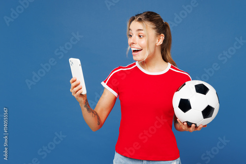 Young shocked surprised woman fan she wear basic red t-shirt cheer up support football sport team hold in hand soccer ball watch tv live stream use mobile cell phone isolated on plain blue background. photo