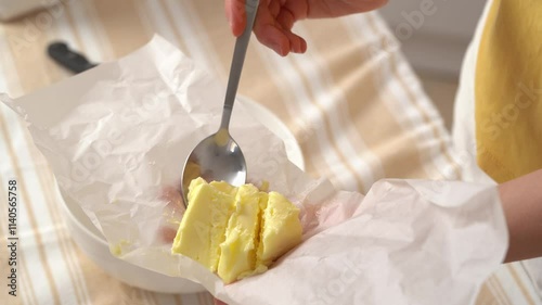 Close-up of a young woman taking spoonful of homemade yellow butter cut from parchment and putting it in bowl for further preparation at kitchen table. Concept of ingredients for dishes, cooking hobby