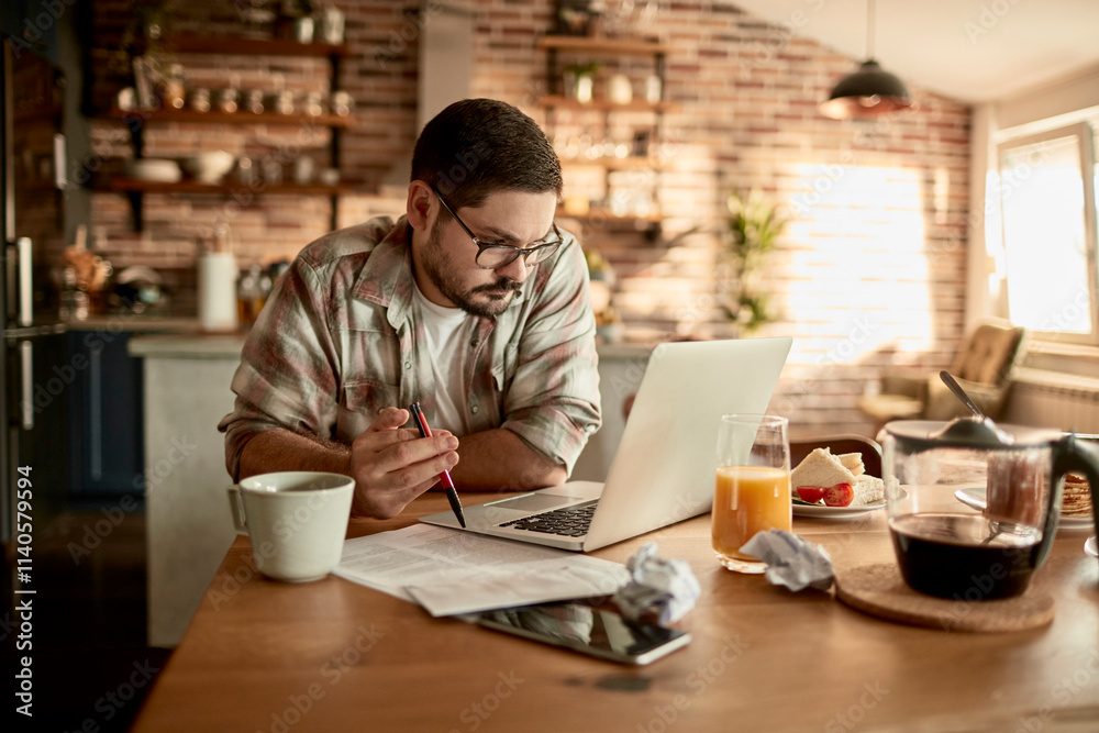 Man working on laptop at kitchen table in a cozy home setting Man working on laptop at kitchen table in a cozy home setting