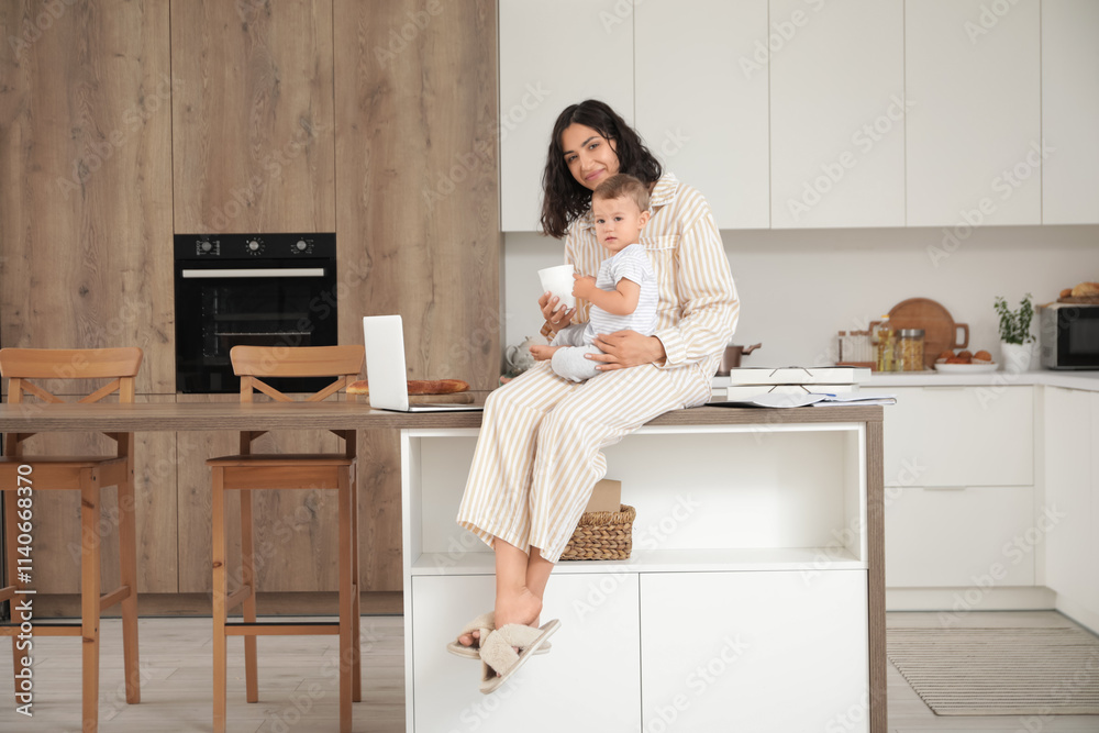 Working mother with coffee cup and her little baby sitting on table in kitchen Working mother with coffee cup and her little baby sitting on table in kitchen