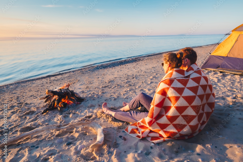 A couple sits wrapped in a blanket on a sandy beach near a campfire, gazing at the calm water and sunset, with a tent visible nearby