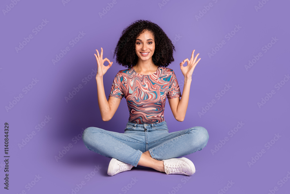 Charming young woman with curly hair sits cross-legged on a violet background, wearing casual jeans and a stylish t-shirt Charming young woman with curly hair sits cross-legged on a violet background, wearing casual jeans and a stylish t-shirt