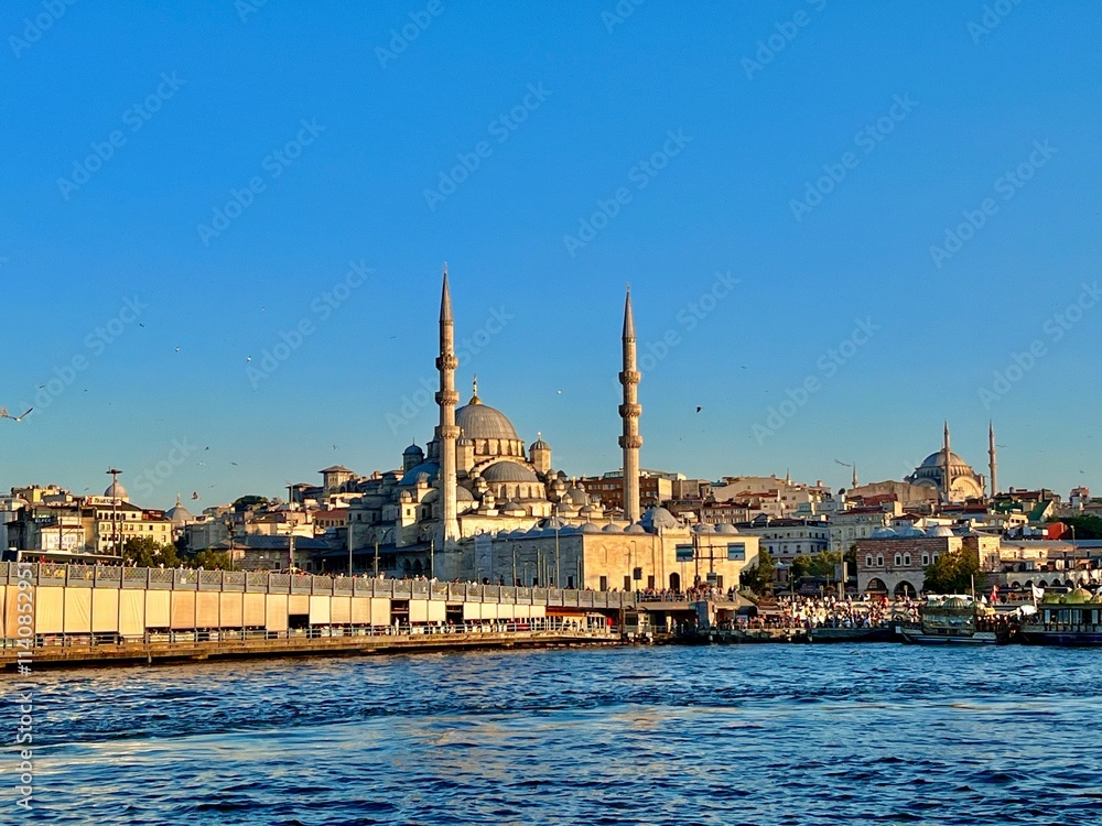 Obraz premium This image shows a scenic view of a waterfront area featuring a prominent mosque with two tall minarets and a large central dome. Surrounding the mosque is a densely built urban area with various 