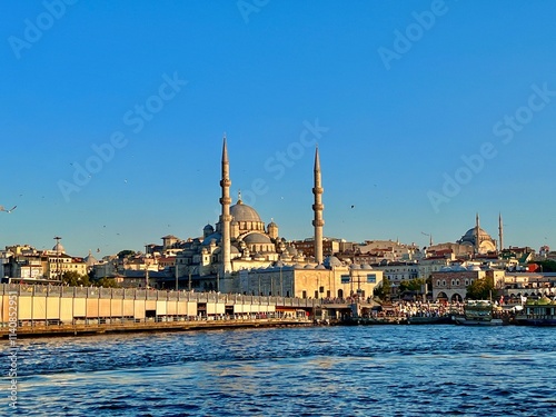 This image shows a scenic view of a waterfront area featuring a prominent mosque with two tall minarets and a large central dome. Surrounding the mosque is a densely built urban area with various 