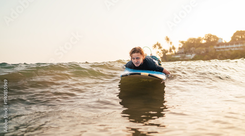 Young man paddling on surfing surfboard on surfing spot in Indian ocean. Palm grove litted sunset rays in the background. Extreme water sports and traveling to exotic countries concept.