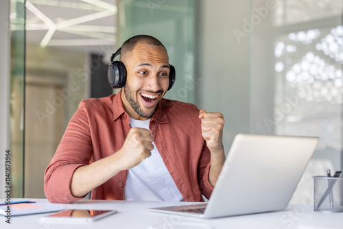 A cheerful man wearing headphones joyfully celebrates an achievement while working on his laptop. Capturing excitement and positive energy in a modern office environment.