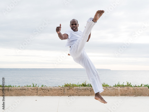 Jumping capoeira martial artist performing a high kick near a scenic ocean backdrop