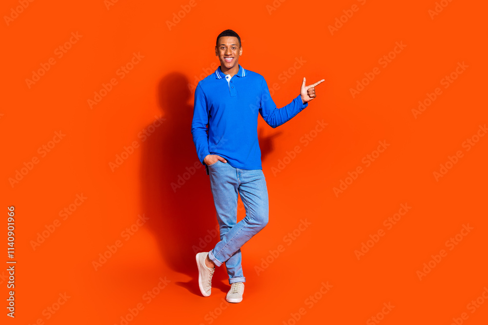 Young man in blue sweater points confidently against orange background Young man in blue sweater points confidently against orange background