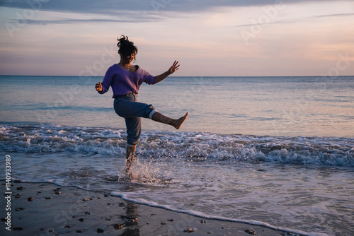 Carefree young woman kicking and splashing ocean waves while enjoying a beautiful sunset at the beach