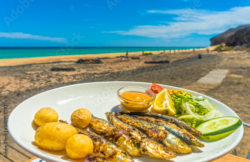 A plate of grilled sardines with salad, potatoes, and sauce is served alongside a refreshing glass of beer at Mal Nombre beach, Fuerteventura, Canary Islands. The bright turquoise ocean and golden san