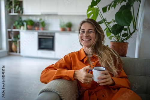 Pleased dreamy middle aged woman sits on couch relax at home holding cup coffee. Joyful female smiling thinking about happy moments, stress free, good mood, wellbeing enjoy weekend time in living room photo