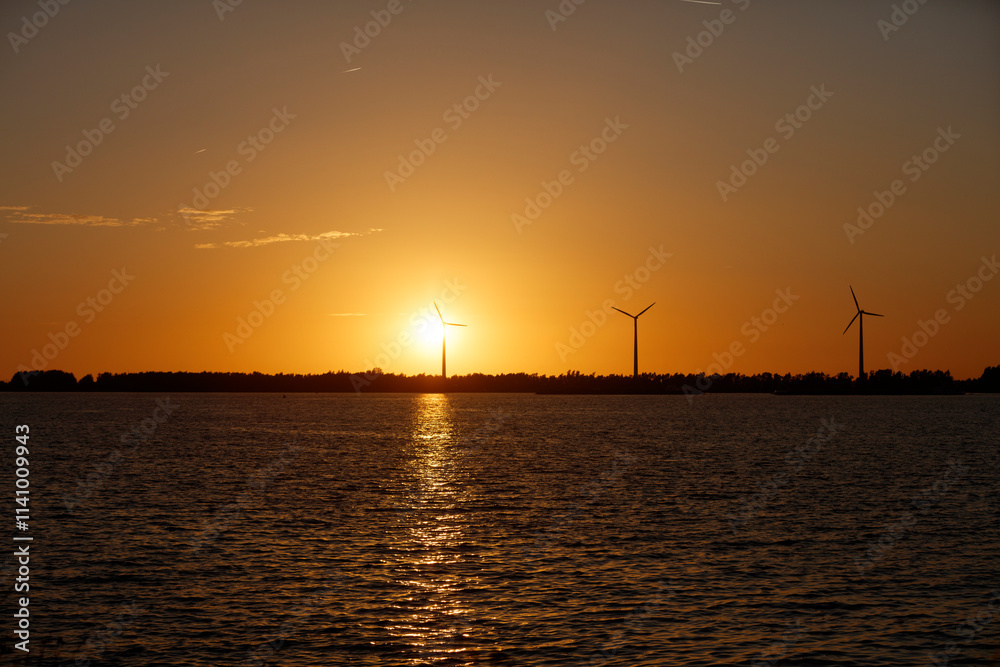 Sunset over water, with wind turbines silhouetted against the orange sky. Calm water reflects the golden light. Sunset over water, with wind turbines silhouetted against the orange sky. Calm water reflects the golden light.