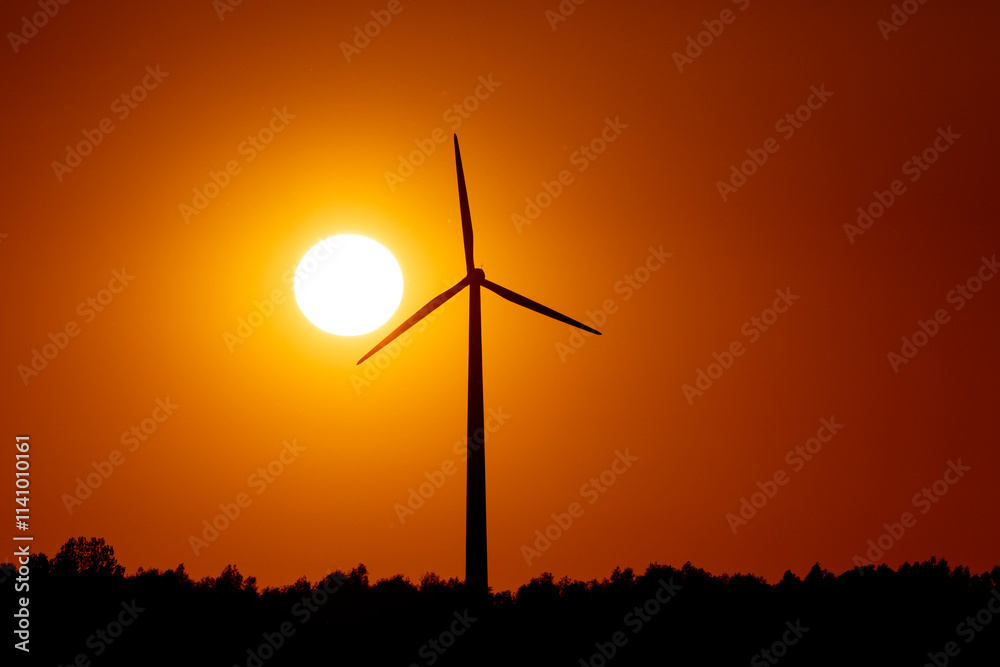 Silhouette of a wind turbine at sunset, with a vibrant orange sky. Trees form a dark line at the horizon. Silhouette of a wind turbine at sunset, with a vibrant orange sky. Trees form a dark line at the horizon.