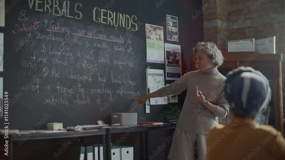 Experienced female teacher standing in sunlit college classroom, pointing at blackboard and explaining grammar rules to students in English class