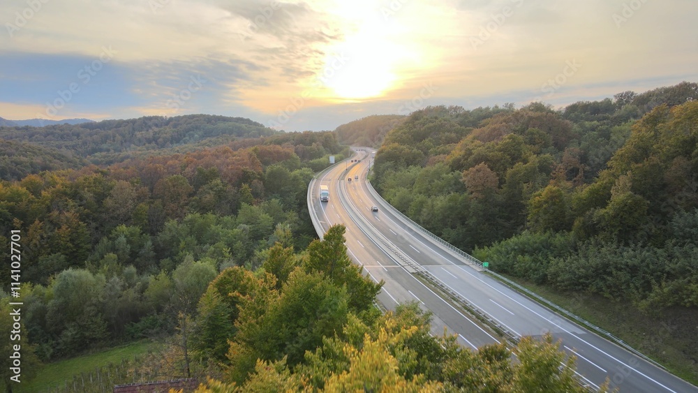 Obraz premium Aerial view of traffic highway through a green forest tree during colorful sunset autumn season. Green energy eco electric car no pollution