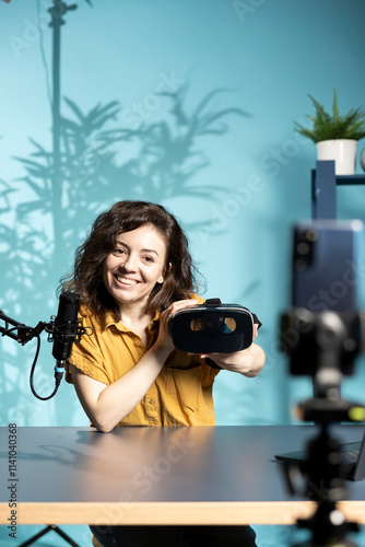 Smiling tech vlogger reviewing VR goggles in living room studio. Happy woman filming virtual reality headset review vlog for gadget enthusiasts using mobile phone and mic