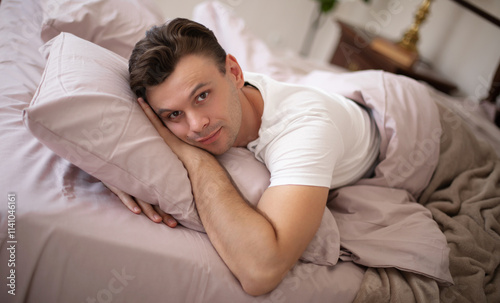 A relaxed man lying on a bed with soft pink bedding, resting his head on a pillow while smiling gently. The cozy bedroom features a warm and serene atmosphere, perfect for relaxation. photo