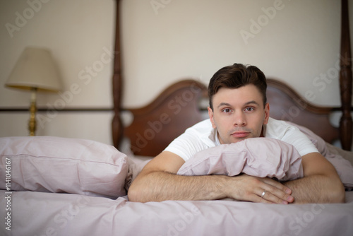 A man lies on a bed with light pink bedding, resting his head on a pillow, gazing forward. The bedroom features classic wooden furniture and a lamp, creating a cozy and elegant atmosphere. photo