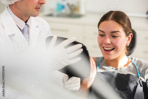 In dentists office, patient sits in dental chair with mirror in hands, see reflection, enjoys result of dental treatment. Male doctor dentist satisfied with result of prosthetics of girl patients